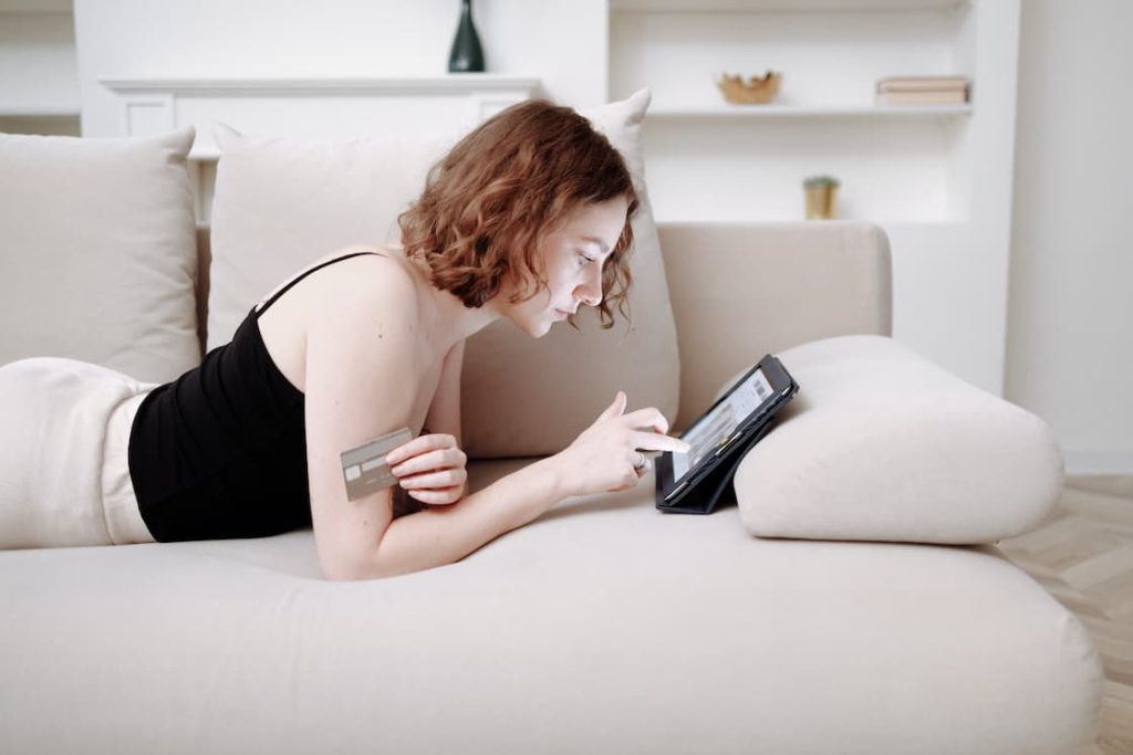 Woman holding a credit card with one hand as she browses through her tablet while lounging on her couch