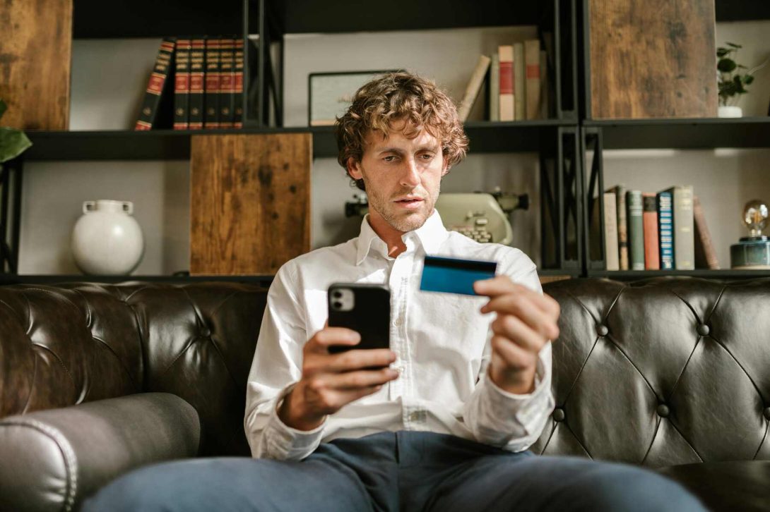 A man sitting on a couch while holding a blue credit card and phone