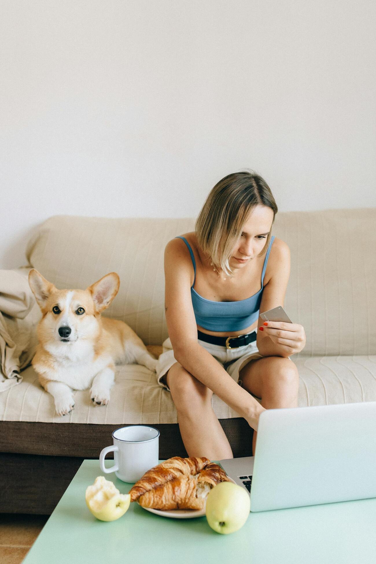 Woman holding a credit card, while sitting on couch with laptop, with a corgi dog beside her