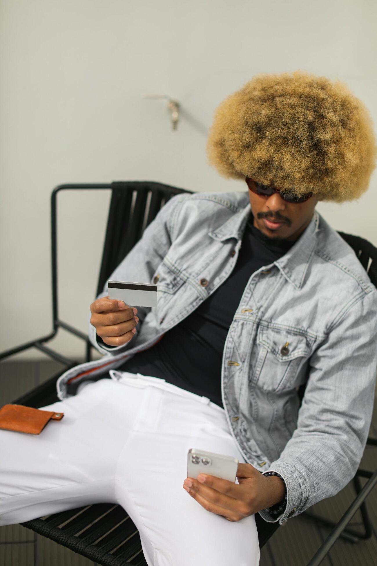 A stylish man with an afro in a denim jacket and white pants holding a phone and credit card