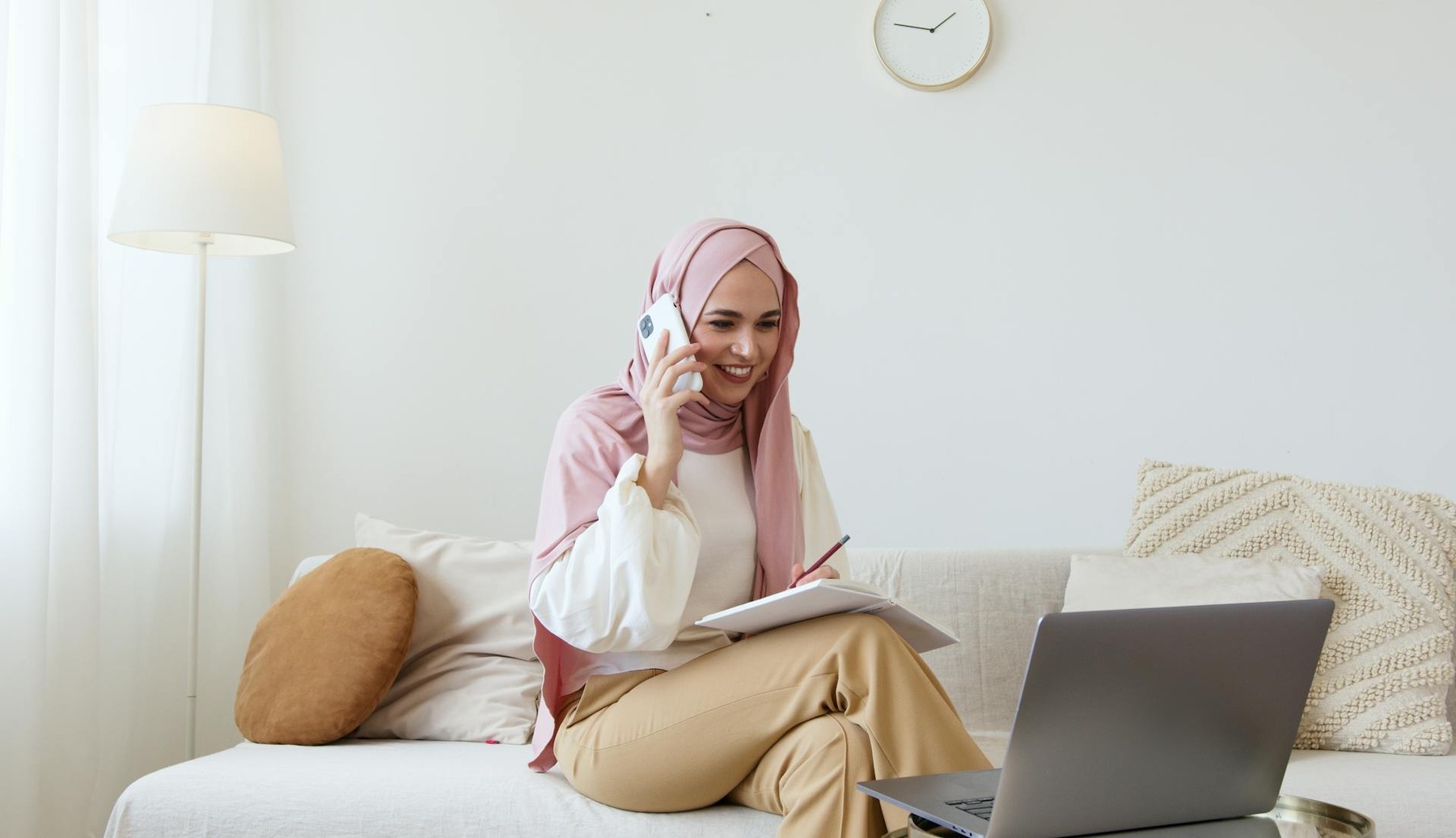 A woman wearing a hijab sitting and talking on a phone in a couch with a laptop and a notebook