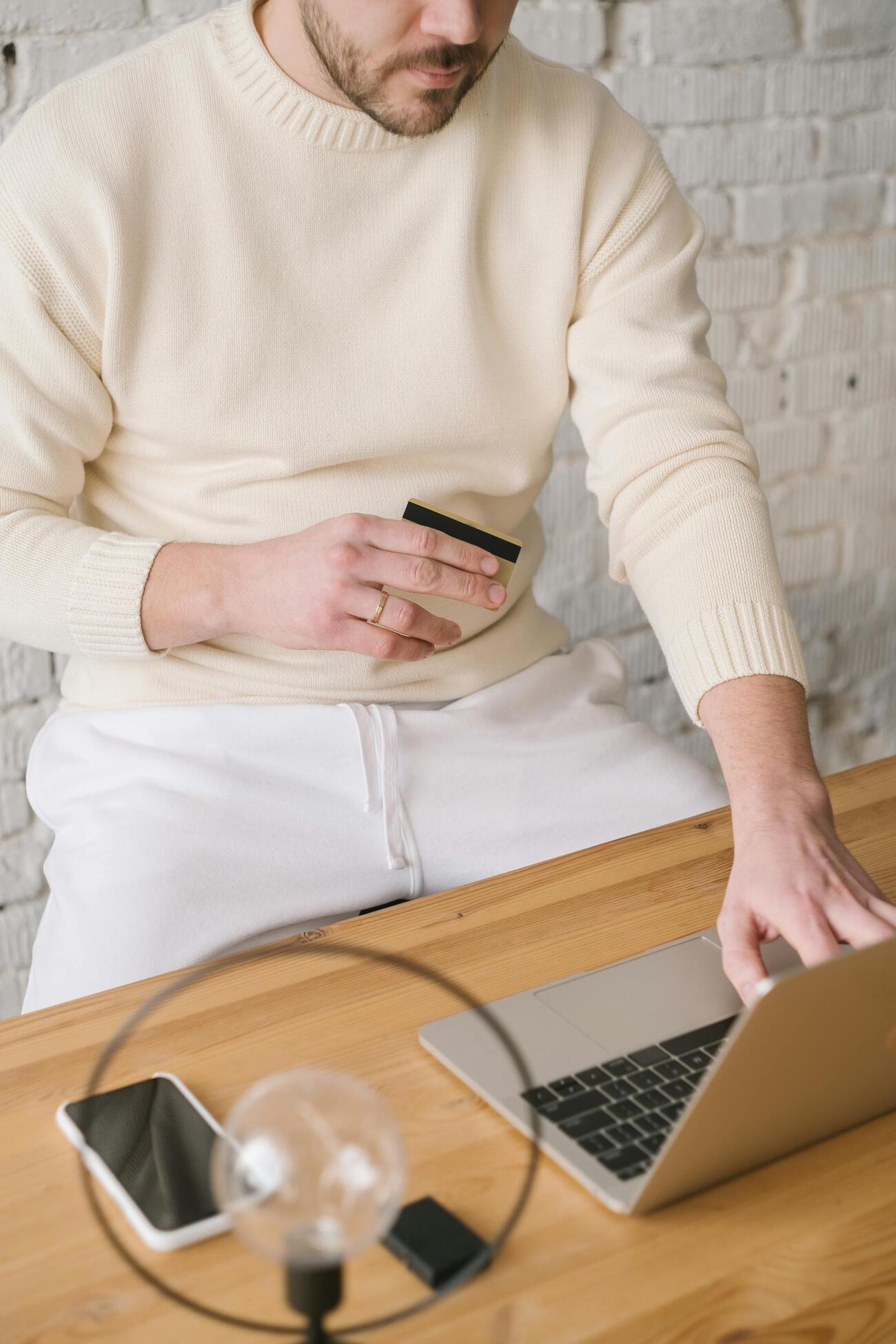 A man in a sweater sitting at a table, using a laptop and holding a credit card