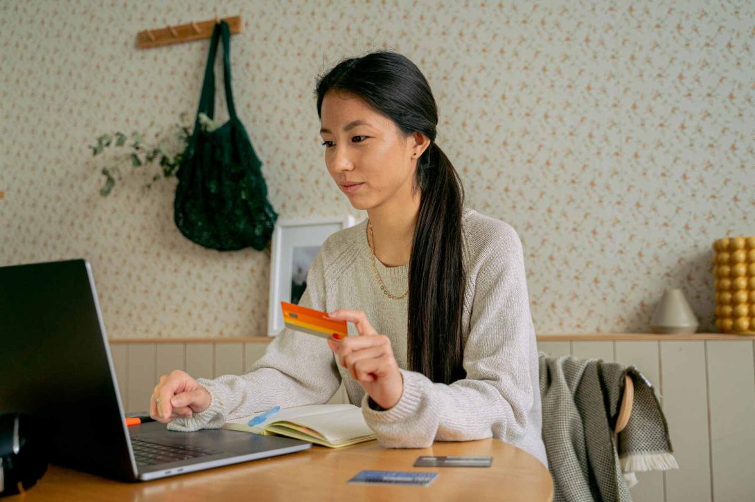 An Asian woman holding an orange credit card while looking at the laptop with tow two more credit cards on the table