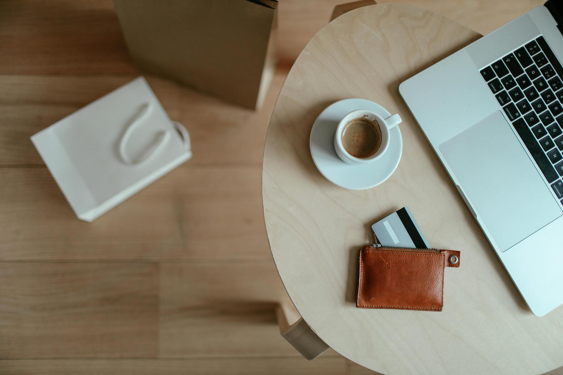 A coffee, wallet with card, and laptop on the table while a shopping bag is on the ground