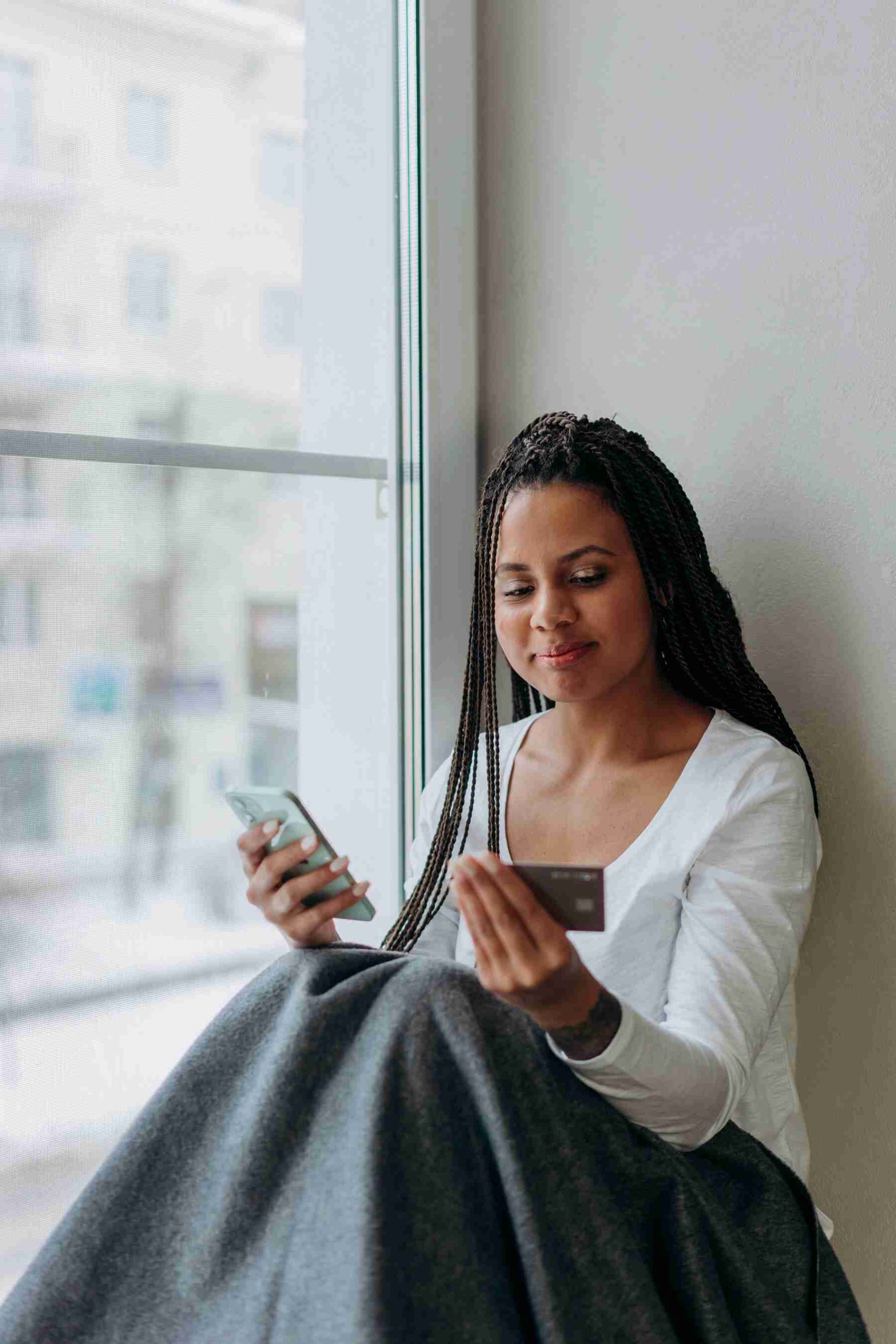 A woman sitting by the window smiling while holding her phone and credit card