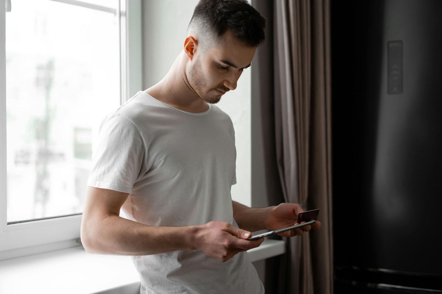 A man standing by the window holding a credit card and a tablet