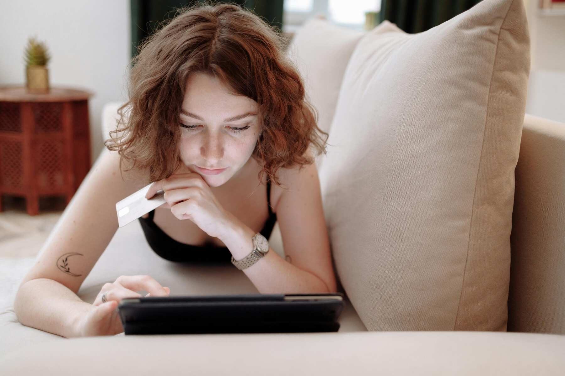 A woman on a couch holding her credit card while browsing on her tablet