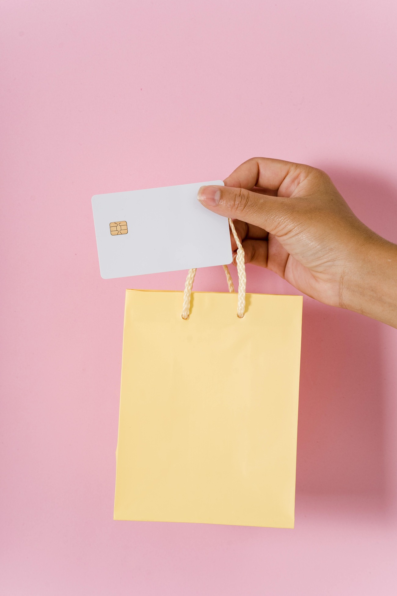 A hand holding a shopping bag with a credit card on a pink background